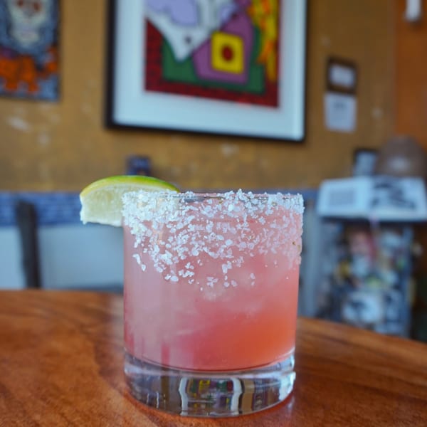 Pink cocktail with a salt rim and lime wedge on a wooden table, with colorful art in the background.