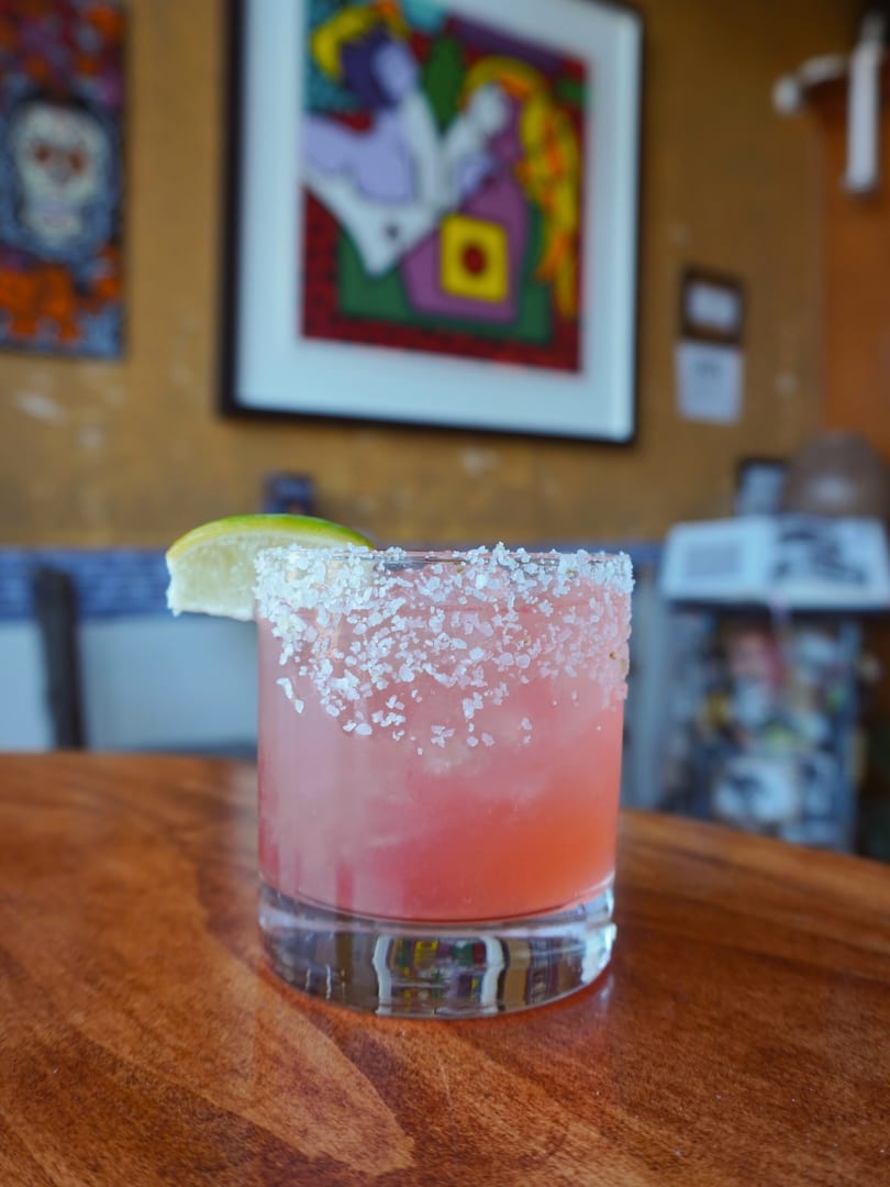 Pink cocktail with a salt rim and lime wedge on a wooden table, with colorful art in the background.