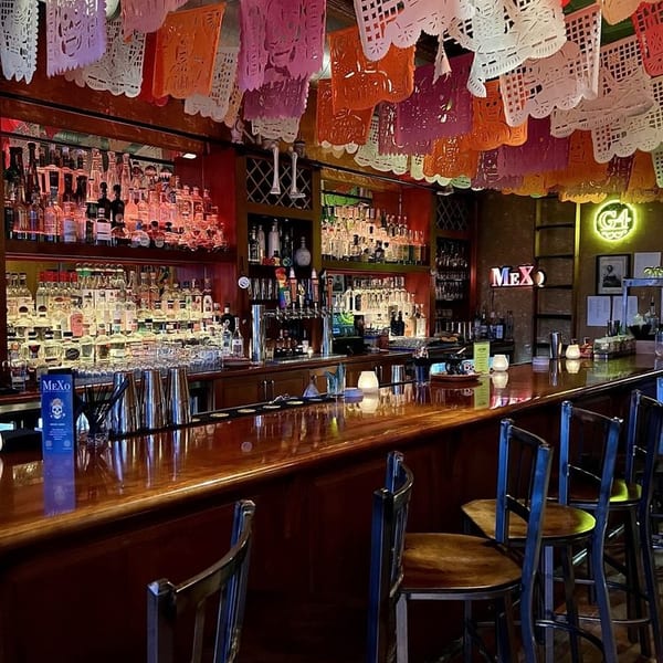 A vibrant bar interior with a long wooden counter, bar stools, and colorful papel picado decorations overhead.