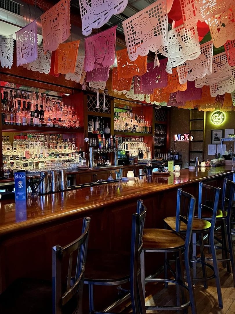 A vibrant bar interior with a long wooden counter, bar stools, and colorful papel picado decorations overhead.