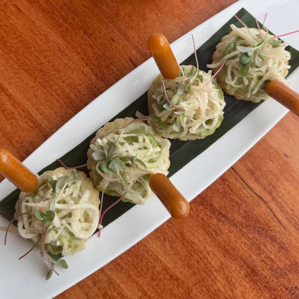 Four savory appetizers with green toppings and small sausages on a white plate with a banana leaf.