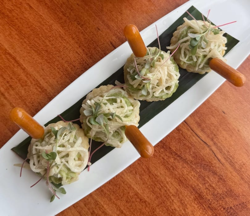 Four savory appetizers with green toppings and small sausages on a white plate with a banana leaf.