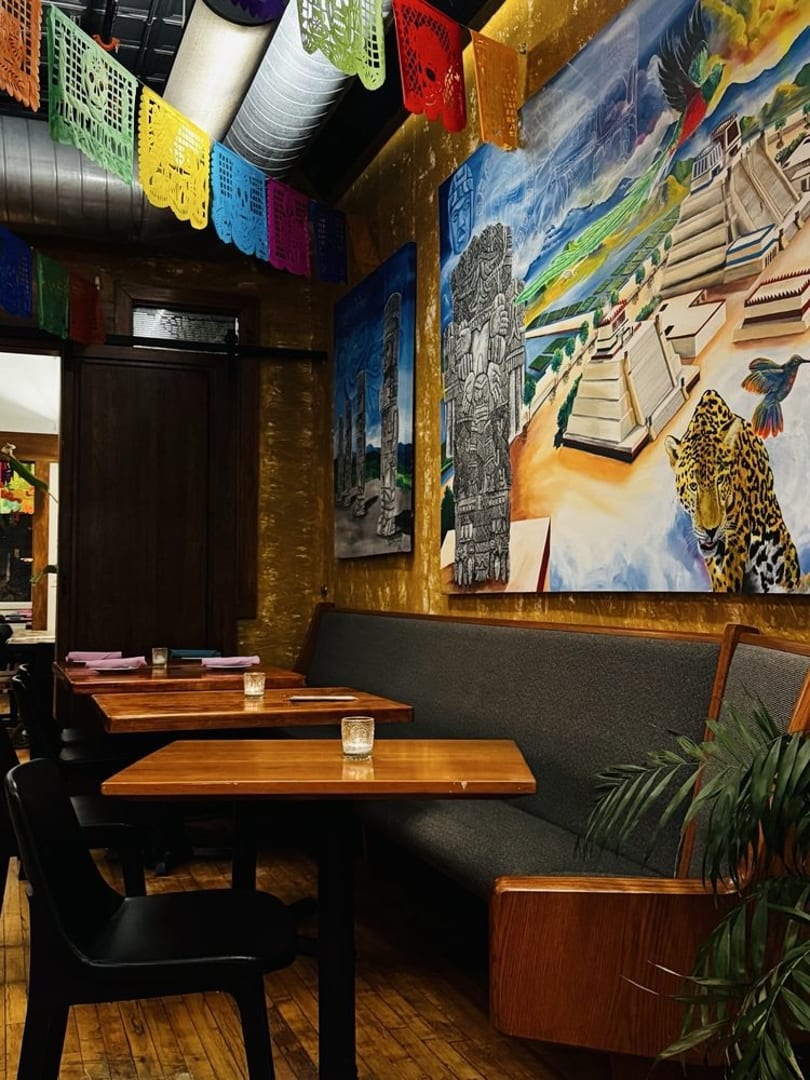 Restaurant interior with colorful papel picado, Mayan-inspired murals, wooden tables, and a long booth.