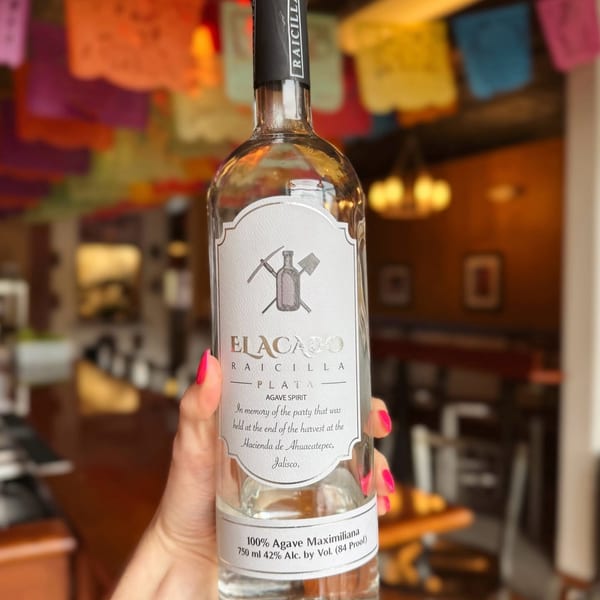 Hand holding a bottle of Elacajo Raicilla agave spirit, with colorful papel picado in the background.
