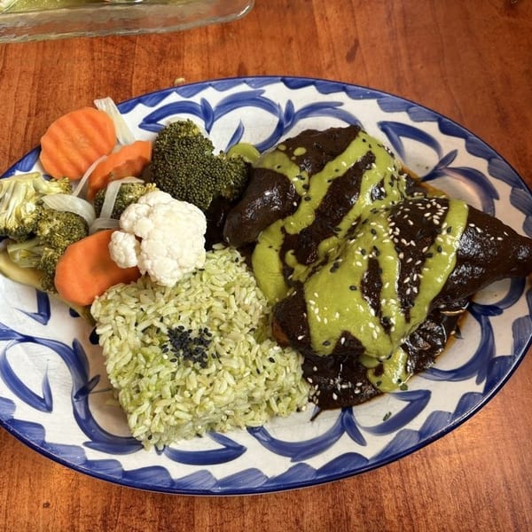 Chicken mole with green sauce, sesame seeds, green rice, and pickled vegetables on a blue and white plate.