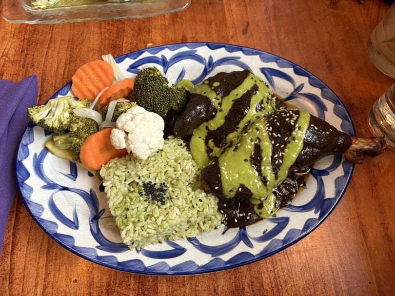Chicken mole with green sauce, sesame seeds, green rice, and pickled vegetables on a blue and white plate.